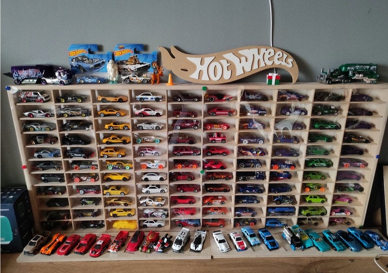 May include: A wooden display shelf with 72 slots for storing Hot Wheels toy cars. The shelf is filled with a variety of colorful cars, including red, blue, green, yellow, and black. The shelf is labeled "Hot Wheels" in white letters on a wooden sign.