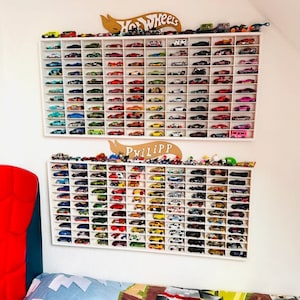 May include: White wall shelves with two rows of white slots holding a collection of toy cars. The top row has a sign that says "Hot Wheels" and the bottom row has a sign that says "Phillip".