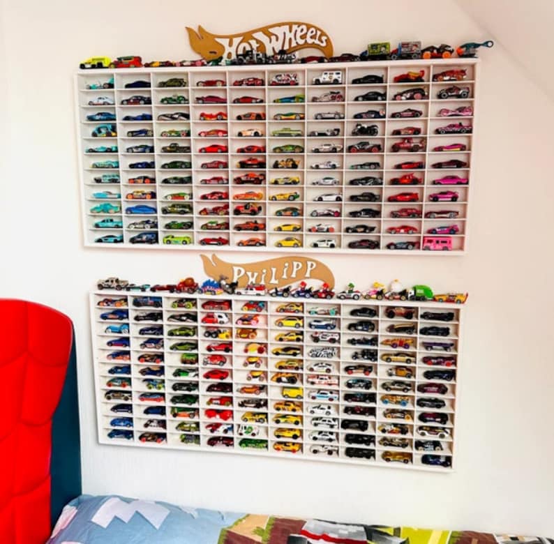 May include: White wall shelves with two rows of white slots holding a collection of toy cars. The top row has a sign that says "Hot Wheels" and the bottom row has a sign that says "Phillip".