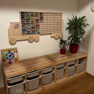 Peut inclure: Une étagère murale en bois en forme de camion jouet avec une grille de compartiments pour ranger des voitures miniatures. L'étagère est fixée à un mur blanc. En dessous de l'étagère, il y a deux étagères en bois avec des bacs de rangement en plastique blanc.