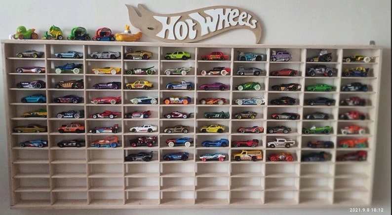May include: A wooden shelf unit with multiple compartments, each holding a small toy car. The cars are in various colors and designs, and the shelf is labeled "Hot Wheels".