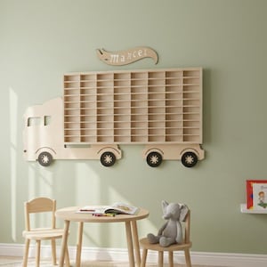 Op de afbeelding: Een houten vrachtwagenvormige plank met meerdere compartimenten, gemonteerd op een lichtgroene muur. De vrachtwagen heeft zwarte wielen en de naam "Marcel" erboven. Een kleine tafel en stoel set staan voor de muur.