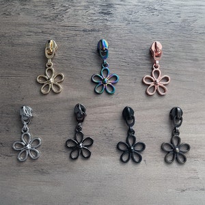 May include: Seven zipper pulls with a flower charm. The charms are in different colors, including gold, silver, black, rose gold, and iridescent.