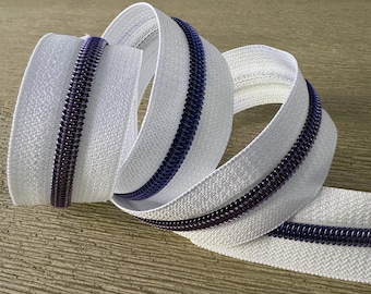 May include: White zipper tape with a purple and blue zipper teeth. The tape is coiled and folded.