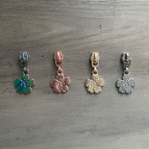 Peut inclure: Quatre tirettes de fermeture éclair en métal avec des breloques en forme de fleur. Les tirettes sont de couleurs irisée, or rose, or et argent. Les breloques en forme de fleur ont des pétales détaillés et sont suspendues à la tirette.