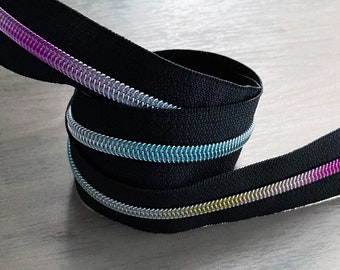 Puede incluir: Primer plano de una cinta de cremallera negra con detalles iridiscentes azules, dorados y morados. La cinta está enrollada y muestra los detalles de los dientes de la cremallera.