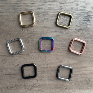 May include: Seven square metal rings in different colors, including gold, silver, black, rose gold, and iridescent. The rings are all the same size and shape, and they are arranged in a grid pattern on a wooden surface.