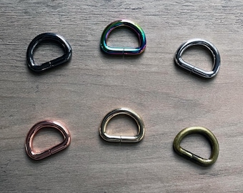May include: Six different metal D-rings in various finishes, including black, silver, rose gold, gold, and iridescent. The rings are arranged in a grid pattern on a wooden surface.