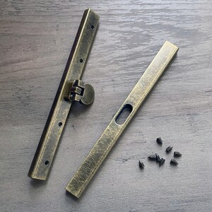 May include: Two antique gold metal clasps with a hinge and small holes for screws. The clasps are on a wooden surface with small black screws scattered around them.
