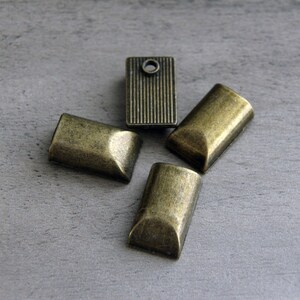May include: Four antique brass-toned metal findings. Three are rectangular with rounded ends, and one is a rectangular shape with vertical lines and a hole. The findings are arranged on a gray surface.