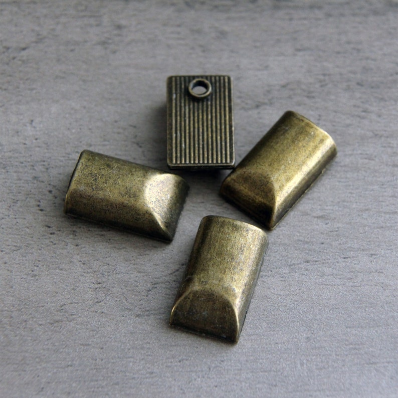 May include: Four antique brass-toned metal findings. Three are rectangular with rounded ends, and one is a rectangular shape with vertical lines and a hole. The findings are arranged on a gray surface.