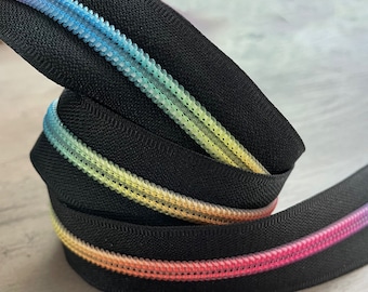 May include: A black zipper with a rainbow gradient teeth. The zipper teeth transition from pink to yellow to blue. The zipper is tightly coiled, showing the texture of the fabric and the colorful teeth.