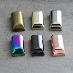 May include: Six rectangular metal end caps in various finishes: antique brass, gold, silver, iridescent, black, and rose gold. Each cap has a rounded bottom edge and a polished, reflective surface, ideal for crafting or jewelry making.