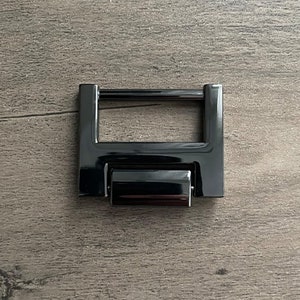 May include: Black metal buckle with a rectangular frame and a sliding mechanism. The buckle is designed for attaching straps or belts.
