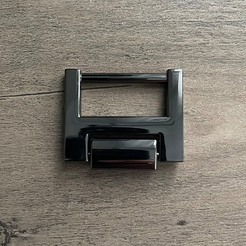 May include: Black metal buckle with a rectangular frame and a sliding mechanism. The buckle is designed for attaching straps or belts.