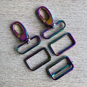 May include: A collection of iridescent rainbow-colored metal hardware components, including two swivel clasps and three rectangular rings. The hardware is displayed on a light-colored, textured surface.