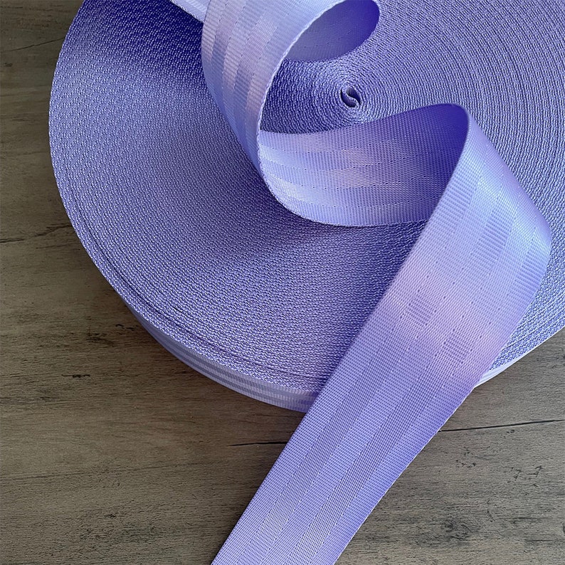 May include: A roll of lavender purple webbing with white stripes. The webbing is rolled up and the end is hanging down.