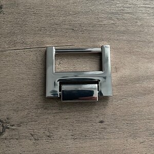 May include: A silver metal buckle with a rectangular frame and a hinged, movable section. The buckle is designed to be used for attaching straps or belts.