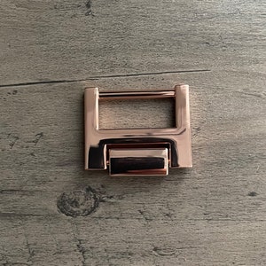 May include: A rose gold metal clasp with a rectangular shape and a sliding mechanism.