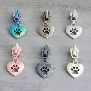 May include: Six zipper pulls with heart-shaped charms featuring a paw print cutout. The charms are in various finishes, including gold, silver, black, rose gold, and iridescent.