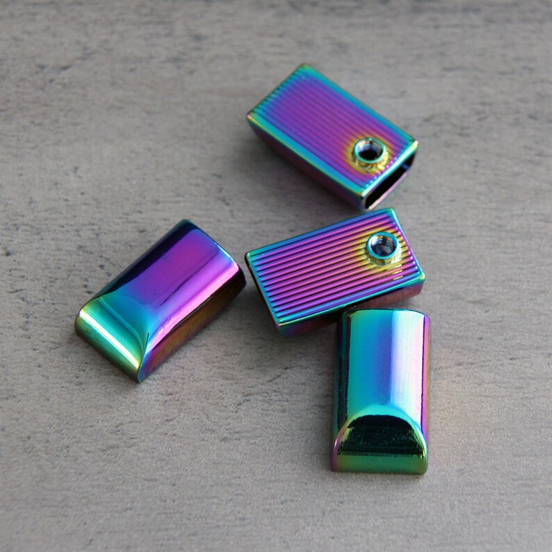 May include: Four rectangular, iridescent metal cord ends. The cord ends have a rainbow finish with shades of purple, blue, green, and yellow. Two of the cord ends have a hole for attaching to a cord. The cord ends are on a gray surface.