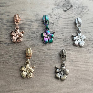 May include: Five zipper pulls with a hibiscus flower charm. The charms are in different colors, including gold, silver, rose gold, and iridescent.