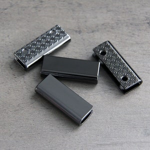 May include: Four black metal slider buckles with a geometric pattern. The buckles are rectangular and have a smooth, shiny finish.