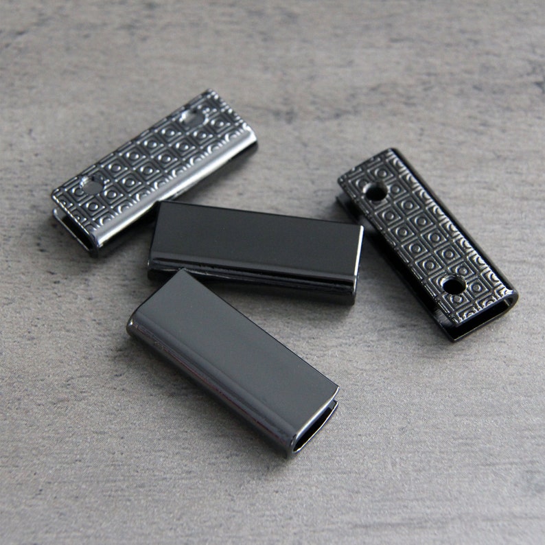 May include: Four black metal slider buckles with a geometric pattern. The buckles are rectangular and have a smooth, shiny finish.