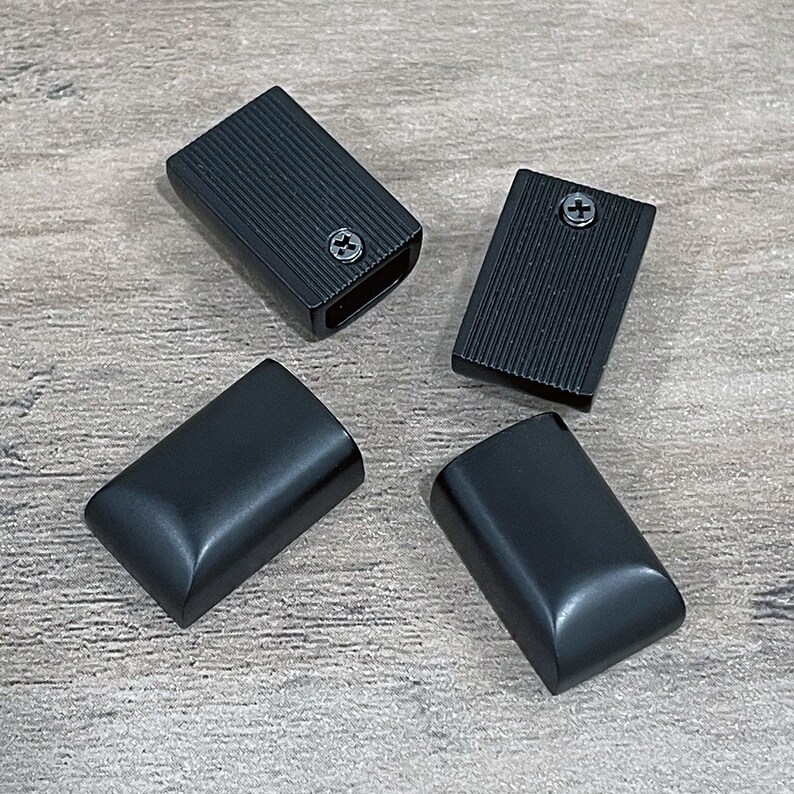 May include: Four black rectangular objects. Two have a ridged texture and a screw. The other two have a smooth, curved surface. All are evenly spaced on a wooden surface.