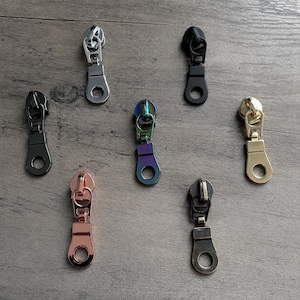 May include: A collection of seven metal zipper pulls in various finishes including silver, matte black, iridescent rainbow, rose gold, antique bronze, and gold. Each pull features a rectangular body with a circular hole at the bottom end.