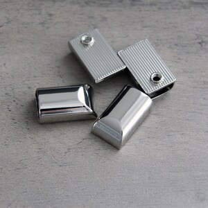 May include: Four shiny, silver-colored metal rectangular clasps. Two clasps have a ridged texture, and a small hole. The other two clasps are smooth and rounded. The clasps are scattered on a gray surface.
