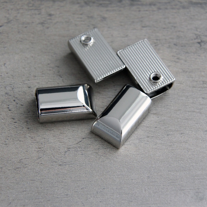 May include: Four shiny, silver-colored metal rectangular clasps. Two clasps have a ridged texture, and a small hole. The other two clasps are smooth and rounded. The clasps are scattered on a gray surface.