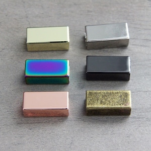 Könnte beinhalten: Sechs verschiedene farbige Metall-Schiebeschließen. Die Schließen sind rechteckig und haben eine glatte Oberfläche. Die Farben sind Gold, Silber, Regenbogen, Schwarz, Roségold und Antikmessing.