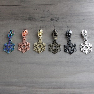 May include: Six snowflake zipper pulls in different colors: iridescent, rose gold, gold, gunmetal, black, and silver.