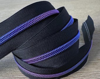 Puede incluir: Primer plano de una cinta de cremallera negra con una bobina iridiscente púrpura y azul. La cinta de cremallera está doblada y apilada sobre una superficie de madera.