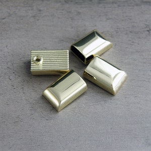 May include: Four gold-colored metal furniture hardware pieces. Three are rectangular with beveled edges, and one is a rectangular block with horizontal grooves and a hole. The pieces are arranged on a gray surface.
