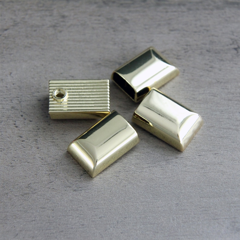May include: Four gold-colored metal furniture hardware pieces. Three are rectangular with beveled edges, and one is a rectangular block with horizontal grooves and a hole. The pieces are arranged on a gray surface.