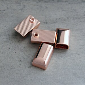May include: Five rose gold rectangular metal beads. Two beads have a ridged surface and a hole at one end. The other three beads have a smooth, curved surface. The beads are scattered on a gray surface.