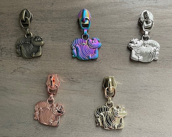 Op de afbeelding: Vijf ritsen met een cartoon Cheshire Cat-ontwerp. De ritsen zijn in verschillende kleuren: goud, rosé goud, zilver, regenboog en zwart.