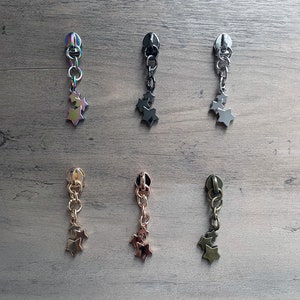 May include: Six zipper pulls with star charms. The zipper pulls are in different finishes: iridescent, black, silver, gold, rose gold, and green.