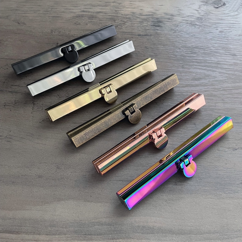 May include: Six different colored metal purse clasps, arranged in a row from left to right. The clasps are black, silver, gold, antique brass, rose gold, and iridescent.