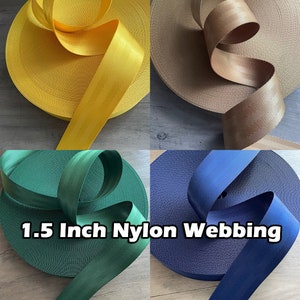 May include: Four spools of 1.5 inch nylon webbing in various colors: yellow, tan, green, and navy blue. The webbing is rolled up, with a portion of each color unrolled to show the texture. The text "1.5 Inch Nylon Webbing" is displayed.