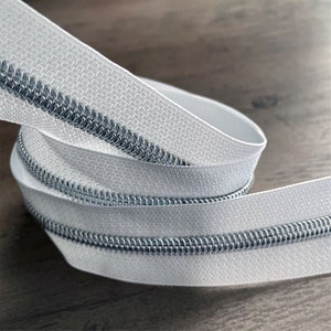 May include: White zipper tape with a silver metal zipper coil. The tape is folded and coiled on a wooden surface.