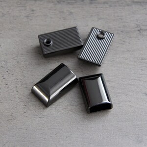 May include: Four rectangular, gunmetal-colored metal findings. Two have a ridged texture, and two are smooth with rounded edges. Each piece has a hole for attaching to other materials. The findings are scattered on a gray surface.