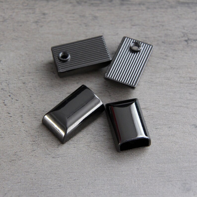 May include: Four rectangular, gunmetal-colored metal findings. Two have a ridged texture, and two are smooth with rounded edges. Each piece has a hole for attaching to other materials. The findings are scattered on a gray surface.