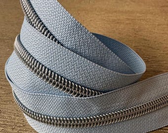 May include: Close-up of a coiled light blue zipper with a silver metal coil. The zipper tape has a textured surface. The zipper is on a wooden surface.