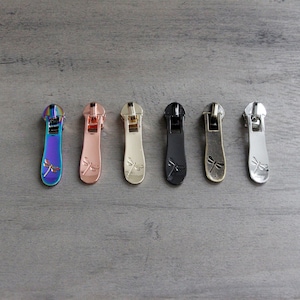 May include: Seven zipper pulls with a dragonfly design. The pulls are in a variety of colors, including rose gold, silver, black, and iridescent.
