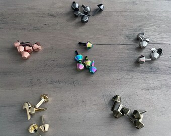 May include: Assortment of metal cone-shaped rivets in various colors, including silver, gold, rose gold, and iridescent. The rivets are arranged in groups on a wood surface.