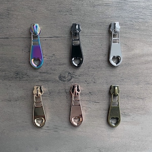 May include: Six zipper pulls with a heart-shaped opening. The pulls are in various colors, including rainbow, black, silver, gold, rose gold, and antique brass.