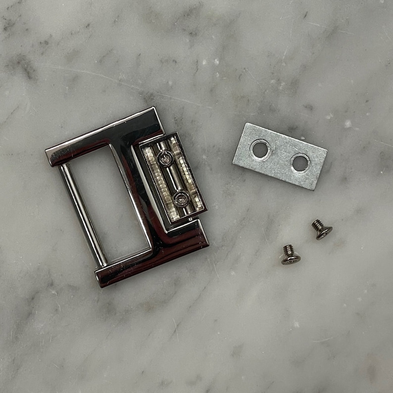 May include: Silver metal buckle with two holes and two screws. The buckle has a rectangular shape with a rounded edge and a small rectangular plate with two holes.